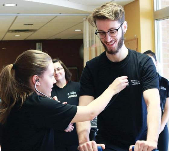 A female Fitness and Health Promotion student checks the  heart-rate of a smiling male student.
