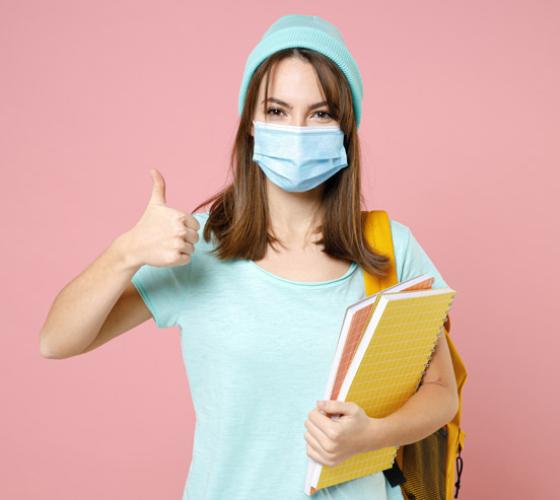 Young woman wearing a mask with a thumb up and holding books.