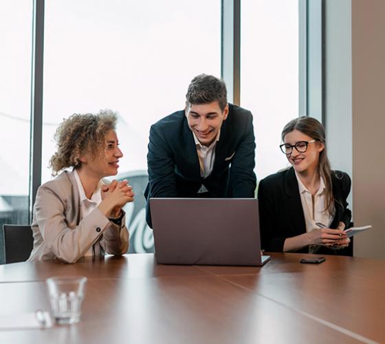 Two women sitting and a man standing in the middle all looking together at a laptop