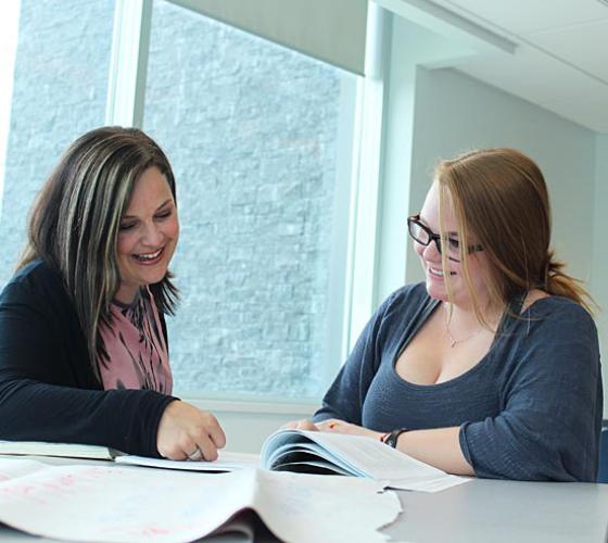 two female students sitting together at a table with books open, discussing and looking at a spot in a book