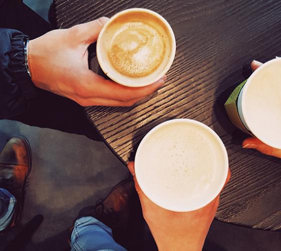 overhead shot of three paper coffee cups filled with coffee sitting on a table. Each person has one hand holding their coffee cup and you can see the floor to some feet