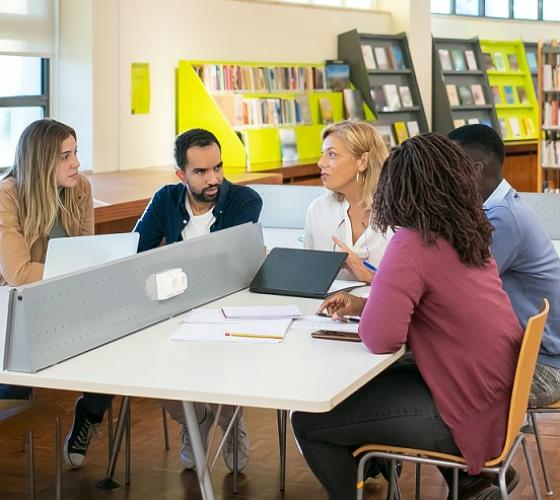 Female instructor sitting at the table with four students listening to her speak in a library setting