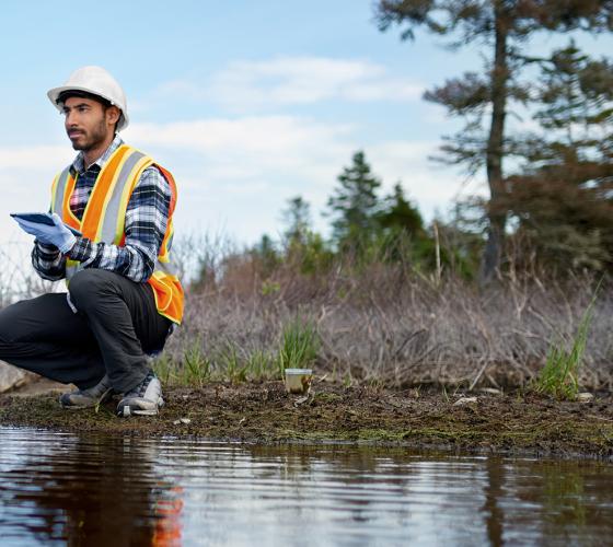 man squatting at edge of river wearing an orange safety vest, white hard hat and white gloves holding a device for studying the area