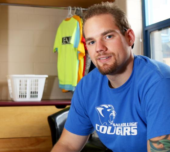 male looking at camera sitting in his residence room with window in background with clothes hanging in open closet and laundry basket