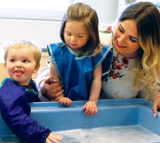 ECE smiling and kneeling down at a water table that three young children are playing at