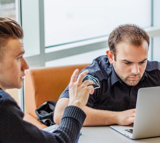 Two Police Foundations students in uniform sitting together at a table one speaking with hands and the other working on a laptop, with lounge chairs and windows in background