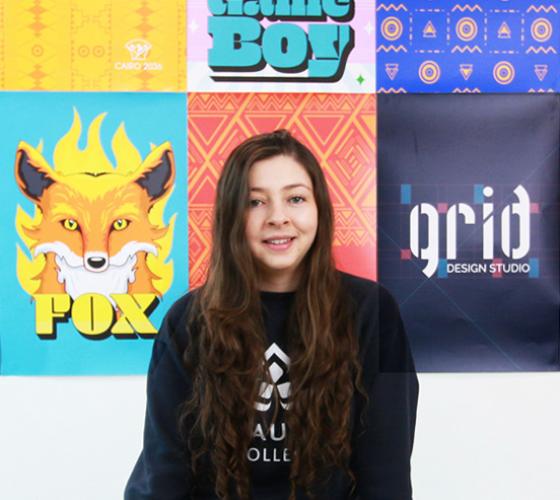 Melissa a new graduate of Graphic Design sitting in front of some of her work displayed on the wall behind her