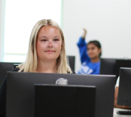 Female student at computer looking up at front of class with students at computers in background