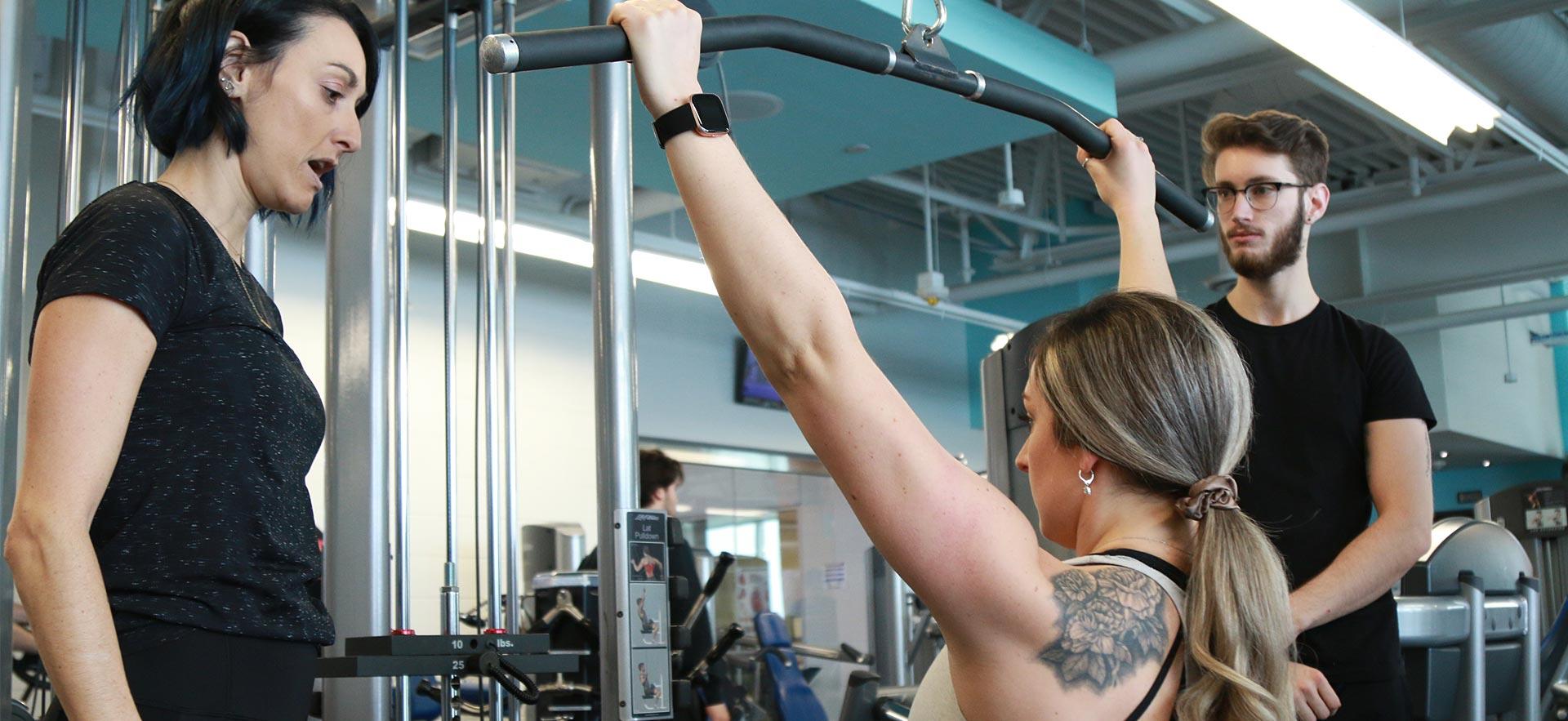 A female fitness and health student is instructed how to use a weight machine.