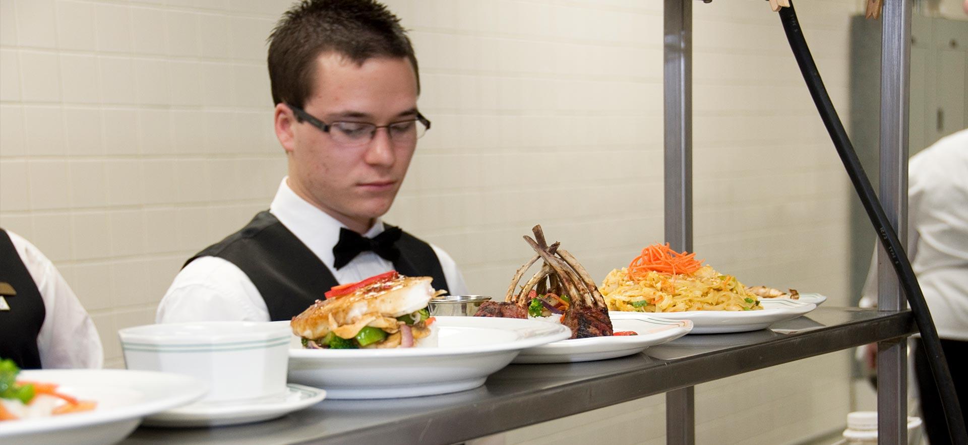 A male Culinary Management student reviews dishes being prepared in one of the culinary kitchens. 
