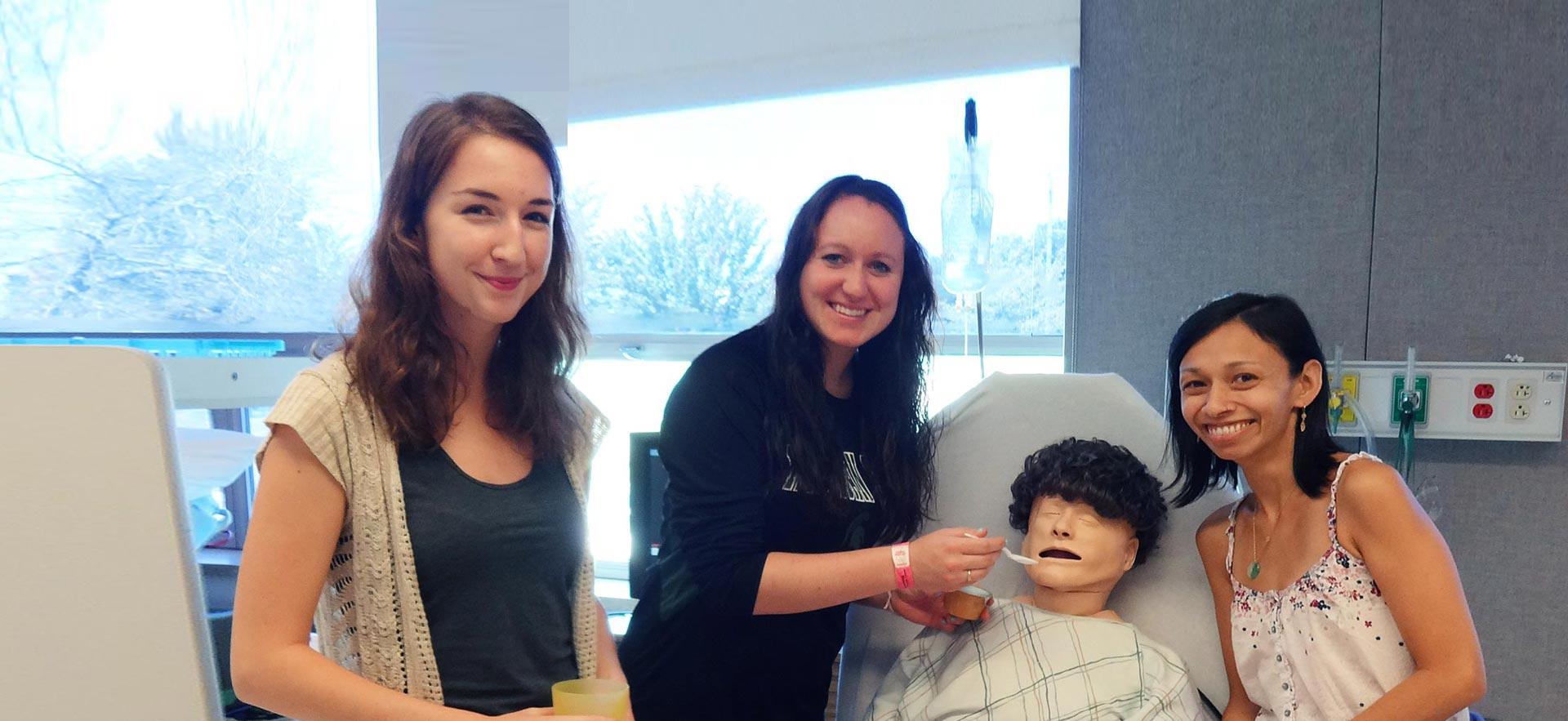 A group of female Personal Support Worker students practice feeding a  healthcare simulation mannequin. 