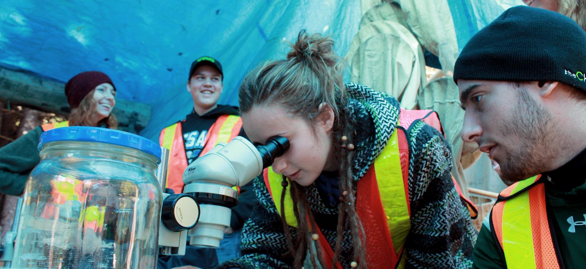 A female  Natural Environment Technician - Conservation and Management student examines a plant sample through a microscope during field camp. 