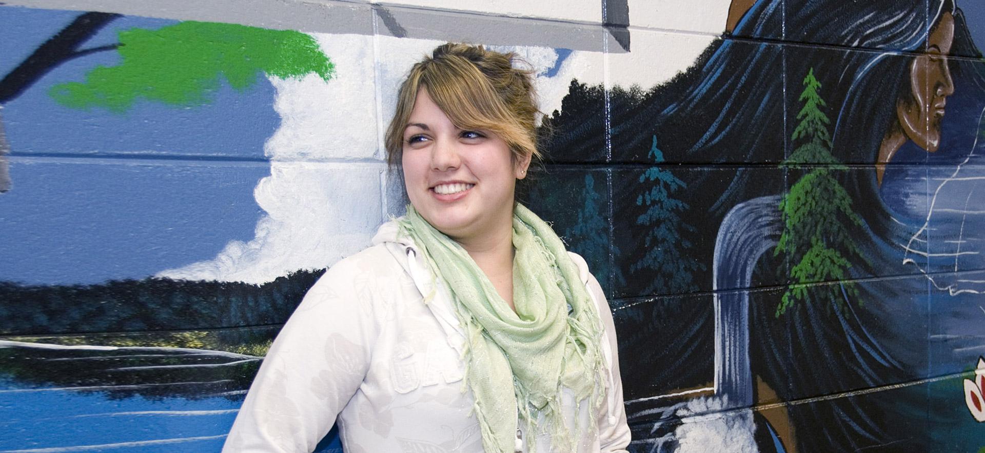 Female indigenous studies student smiling in front of mural.