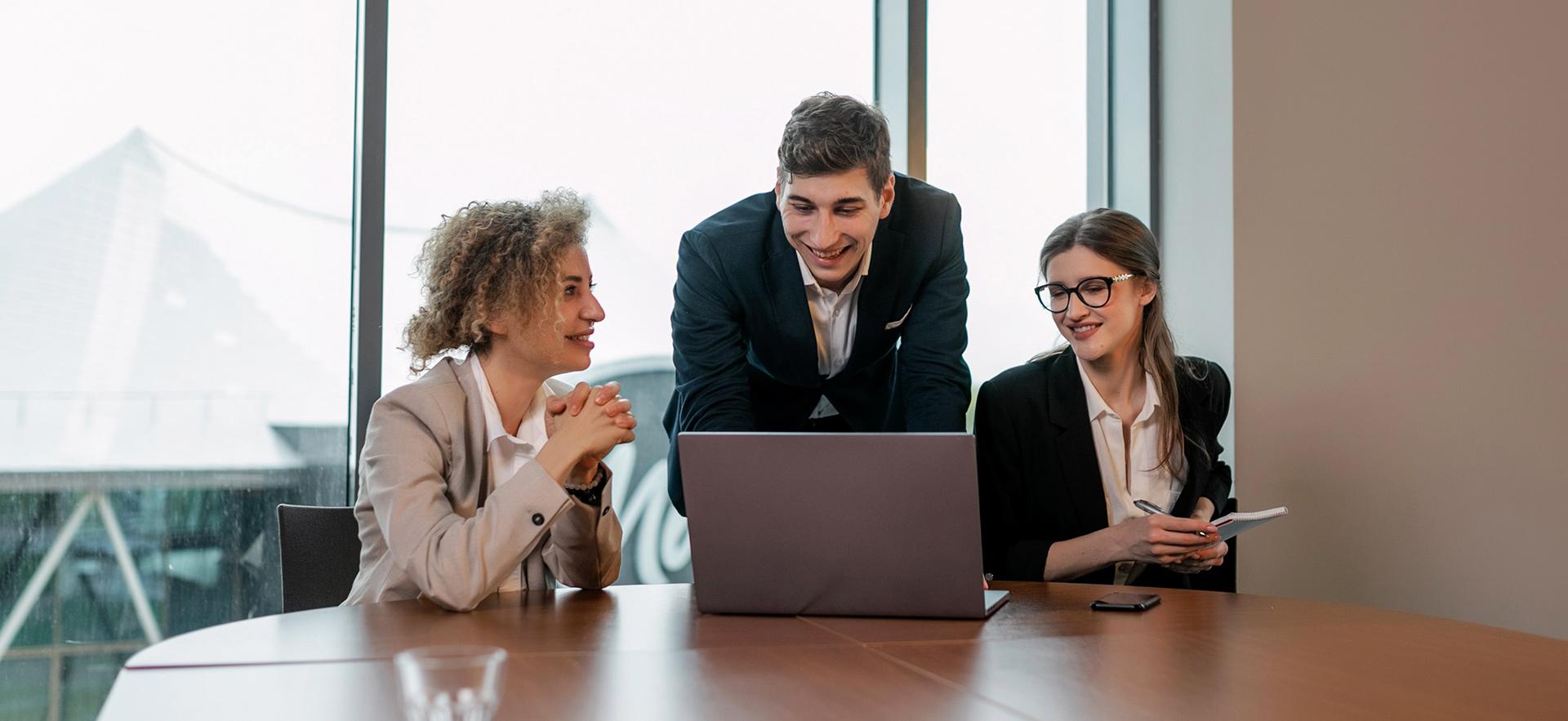 Two women sitting at a desk and a man standing in the middle all looking together at a laptop