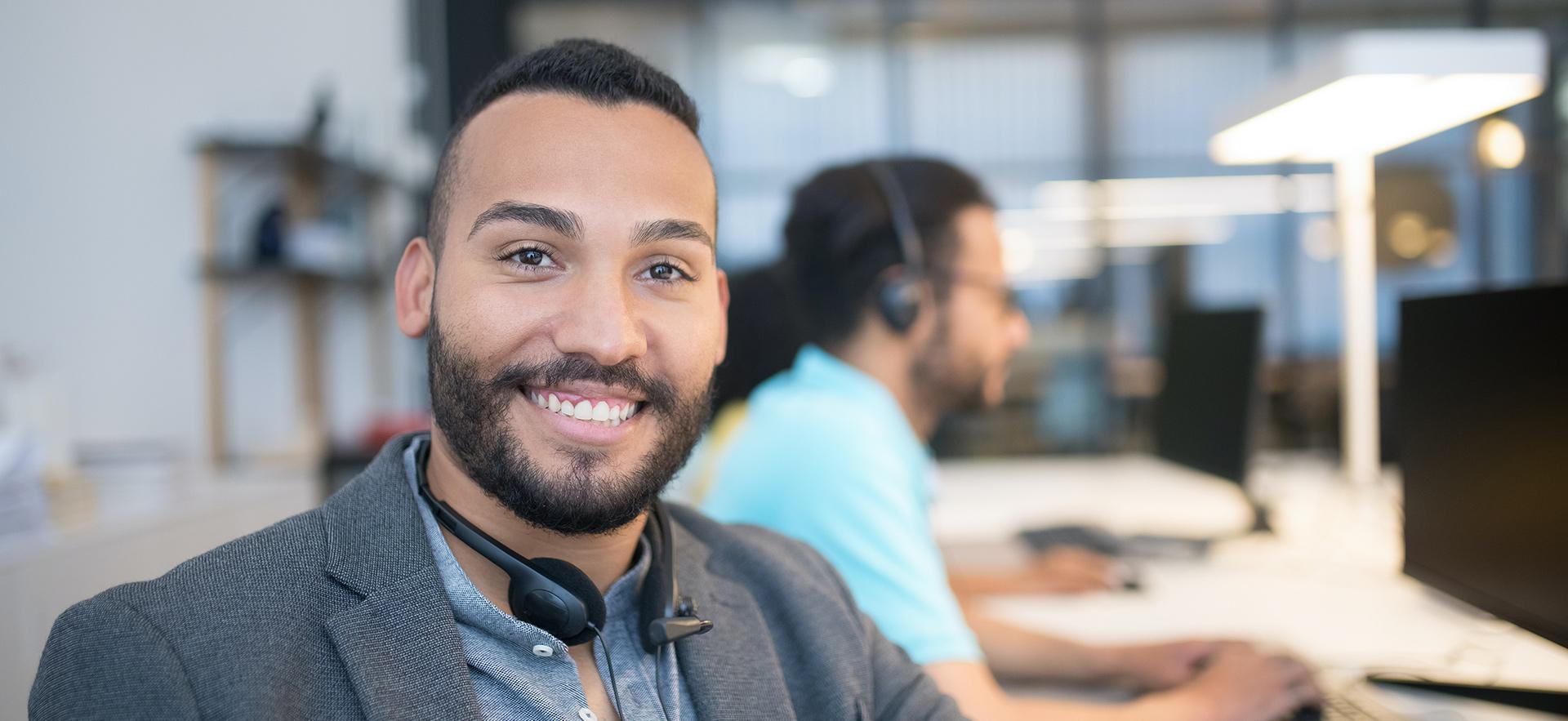 Man smiling at desk with headset around neck and another man in background in large office setting