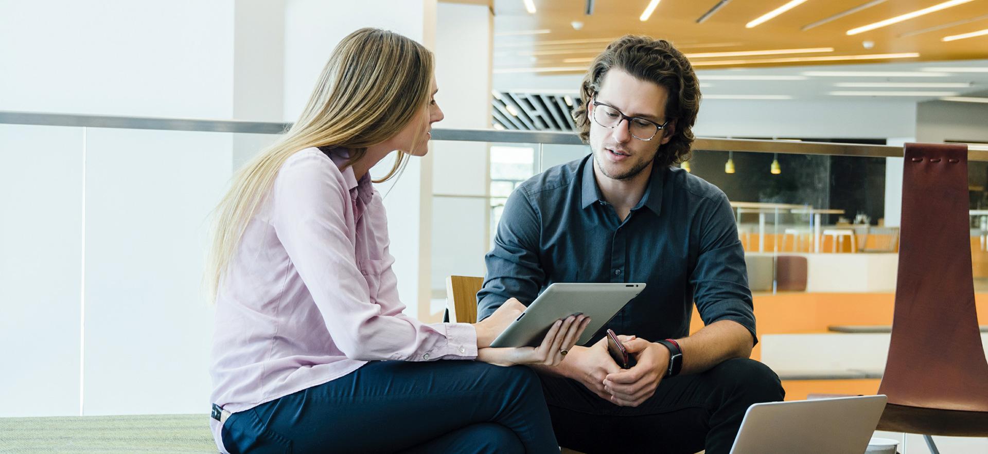 Man and woman sitting in an open office area looking at a tablet together and talking