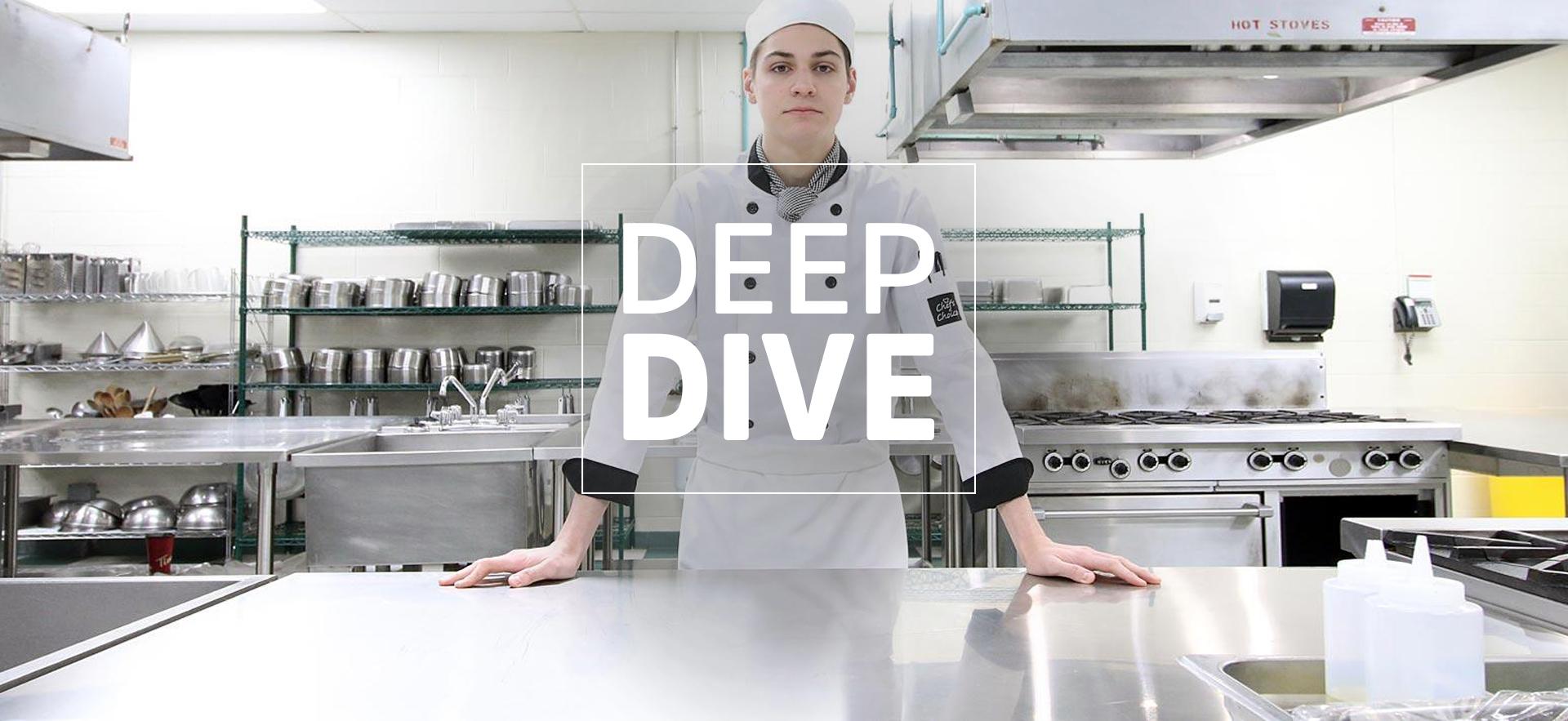 culinary student wearing his chef uniform standing at a stainless steel counter with hands on it and looking at camera with kitchen in background