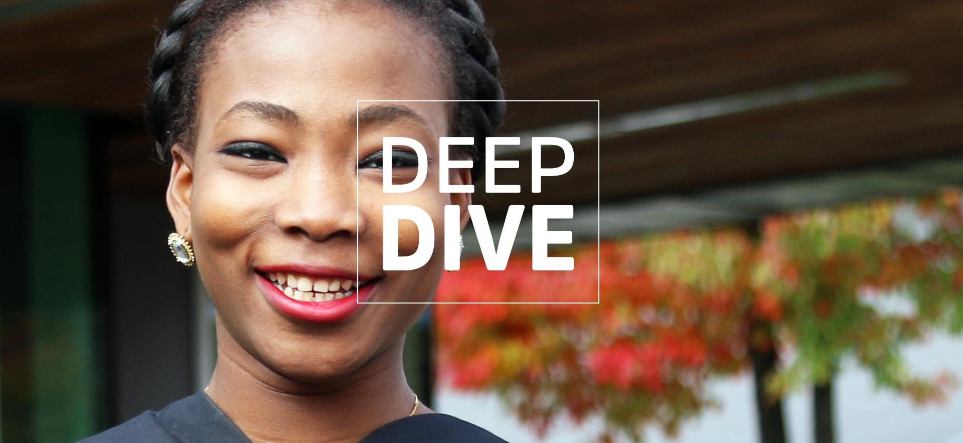graduate in robe shown neck up smiling at camera with tree seen through window in background and white text Deep Dive overlay 