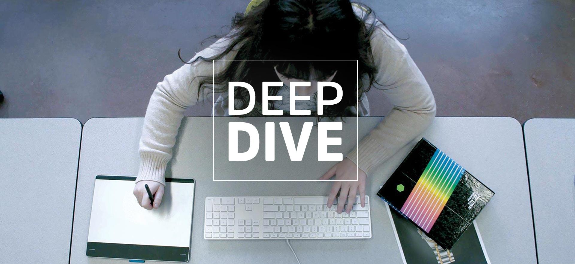 Student shown overhead while sitting at desk with one hand on a computer keyboard and the other with a drawing pen in hand drawing on a tablet and printout and book on the opposite side of her