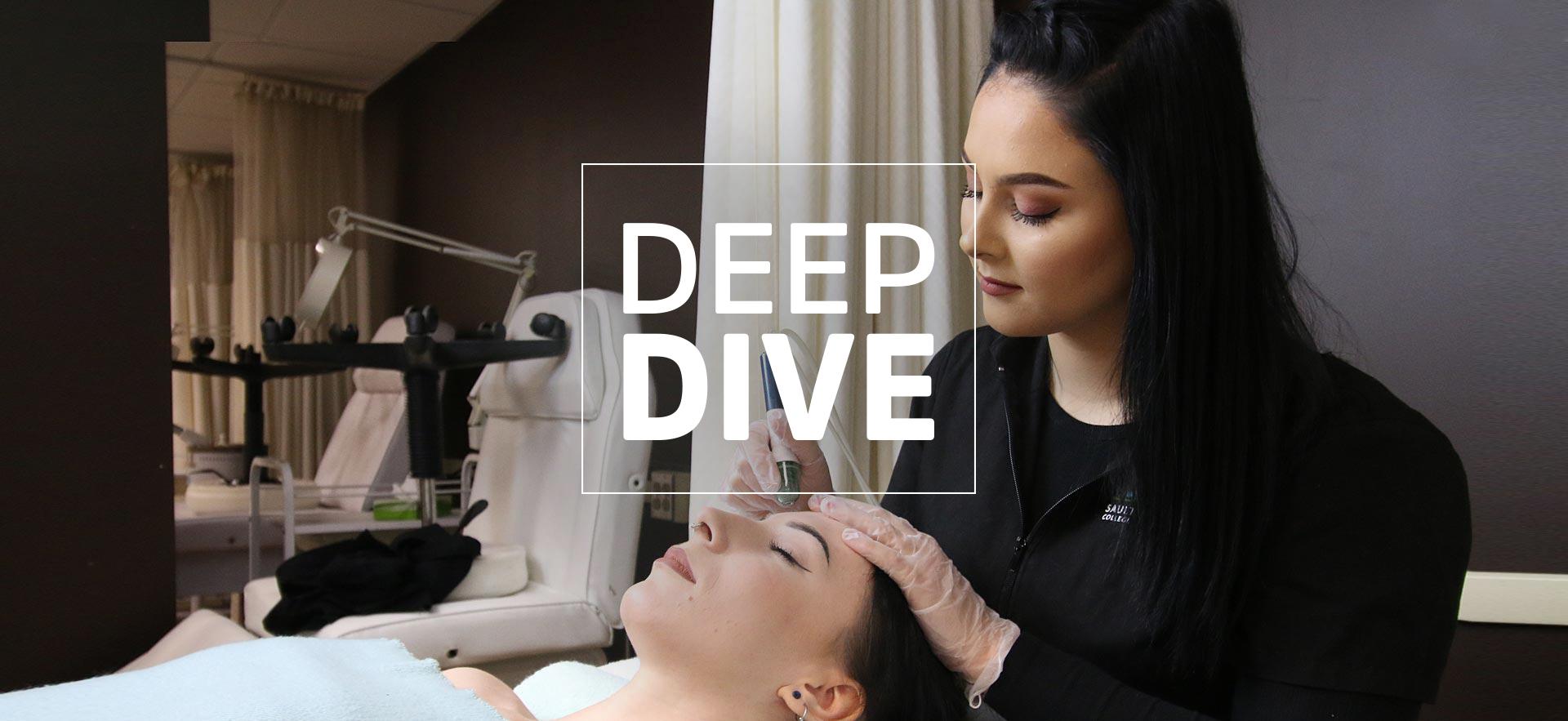 Esthetician student in uniform sitting at the back of a client's head while she lays on salon table with eyes closed to receive a facial treatment