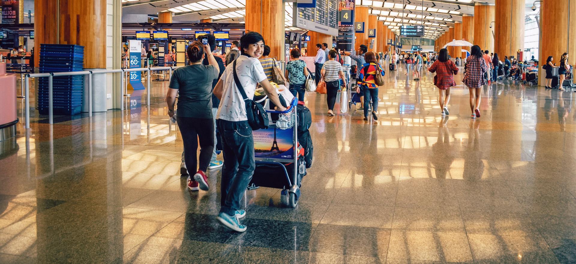 inside an airport showing people walking around and one young adult pushing a luggage cart and looking back smiling at the camera