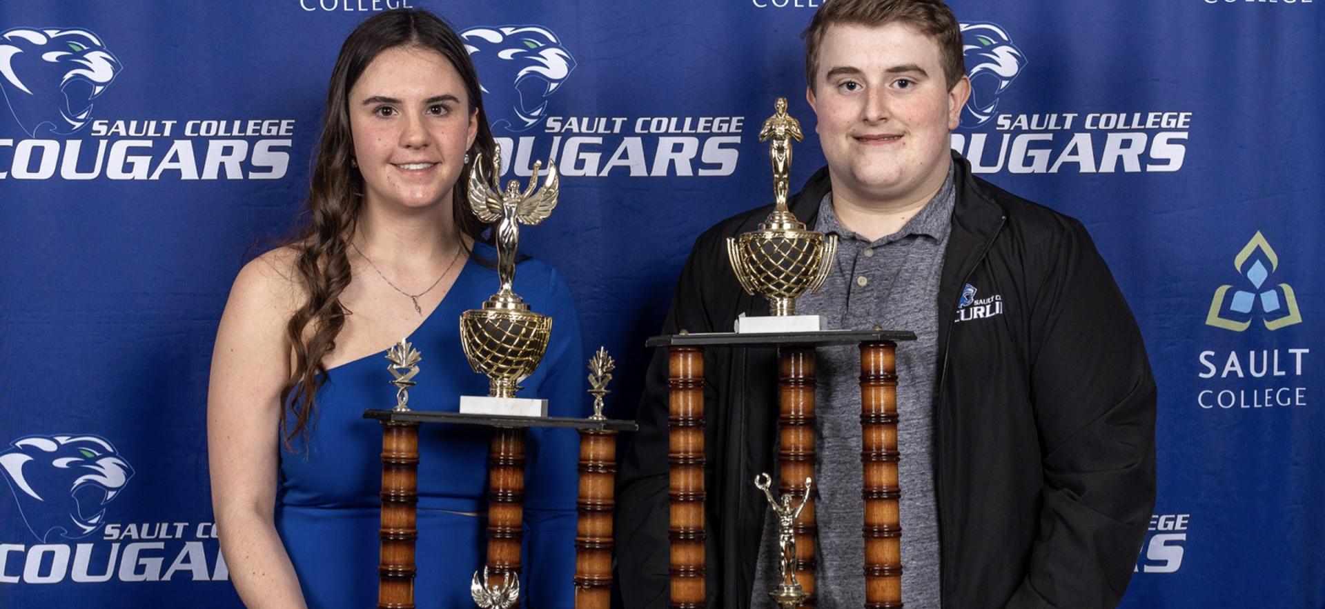 women in blue dress and man in black coat and grey shirt hold trophies