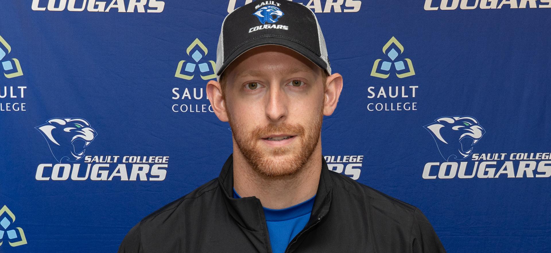 man with beard Sault College cougars black ball cap and jacket standing in front of Sault College cougars backdrop