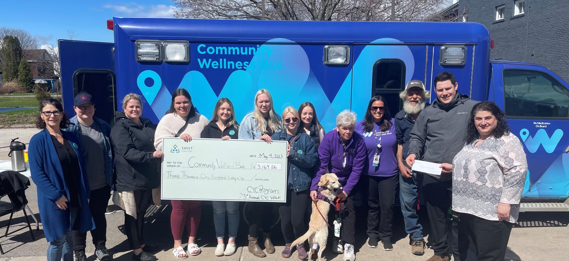 13 people standing and one dog (sitting) in front of a dark blue and light blue bus. Three of the people are holding a large cheque made out to the Community Wellness Bus for $3196.