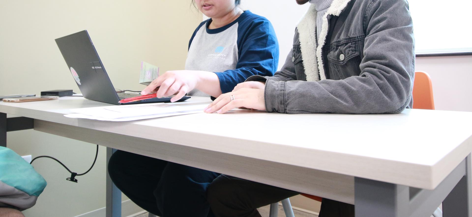 women sitting at table in white shirt with blue sleeves in front of a laptop computer. Sitting beside her is a male with a baseball cap and grey sweater