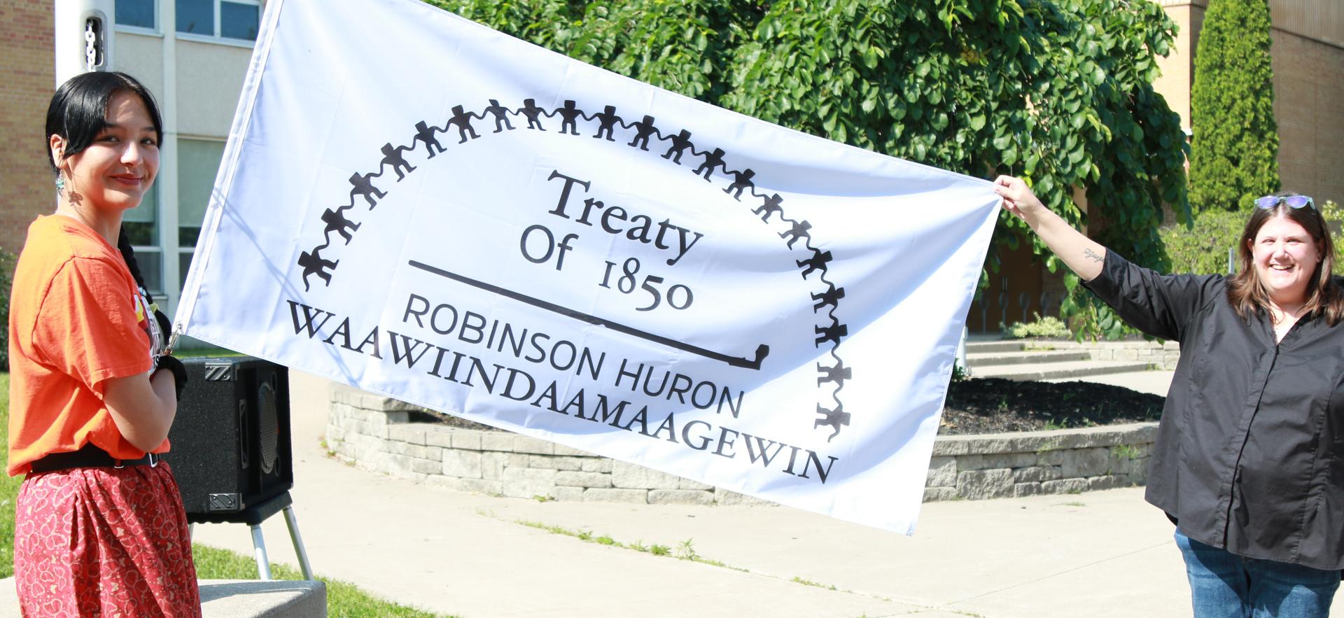 woman in orange shirt on the left hold up Robinson Huron Treaty flag with woman on right in black shirt