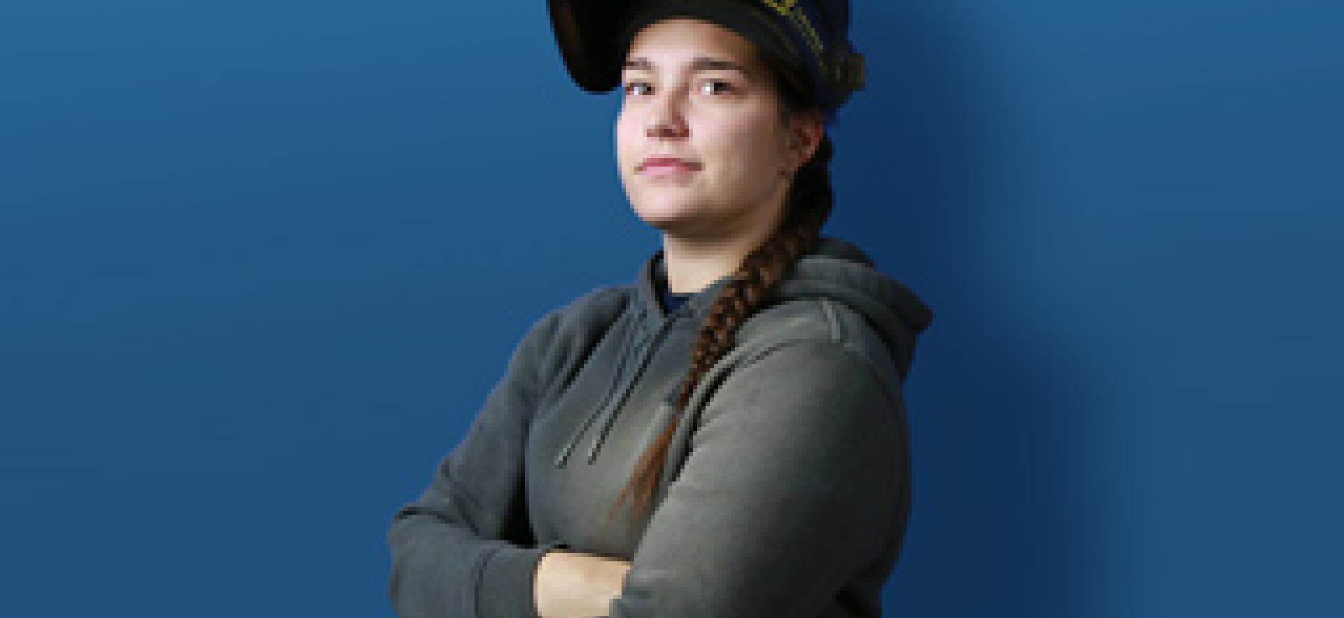 Skilled Trades student wearing a welding mask lifted up off her face standing in front of blue background with text YOUR TIME TO BE SKILLED