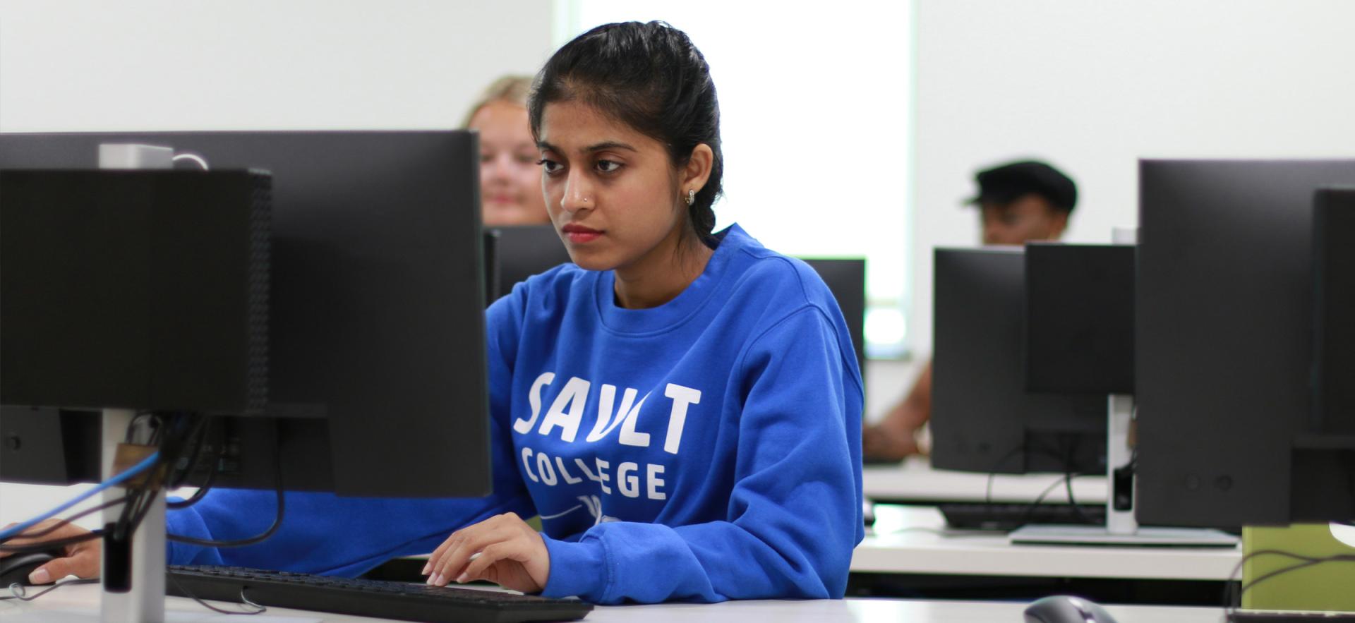 Student sitting at computer in class wearing blue Sault College sweater