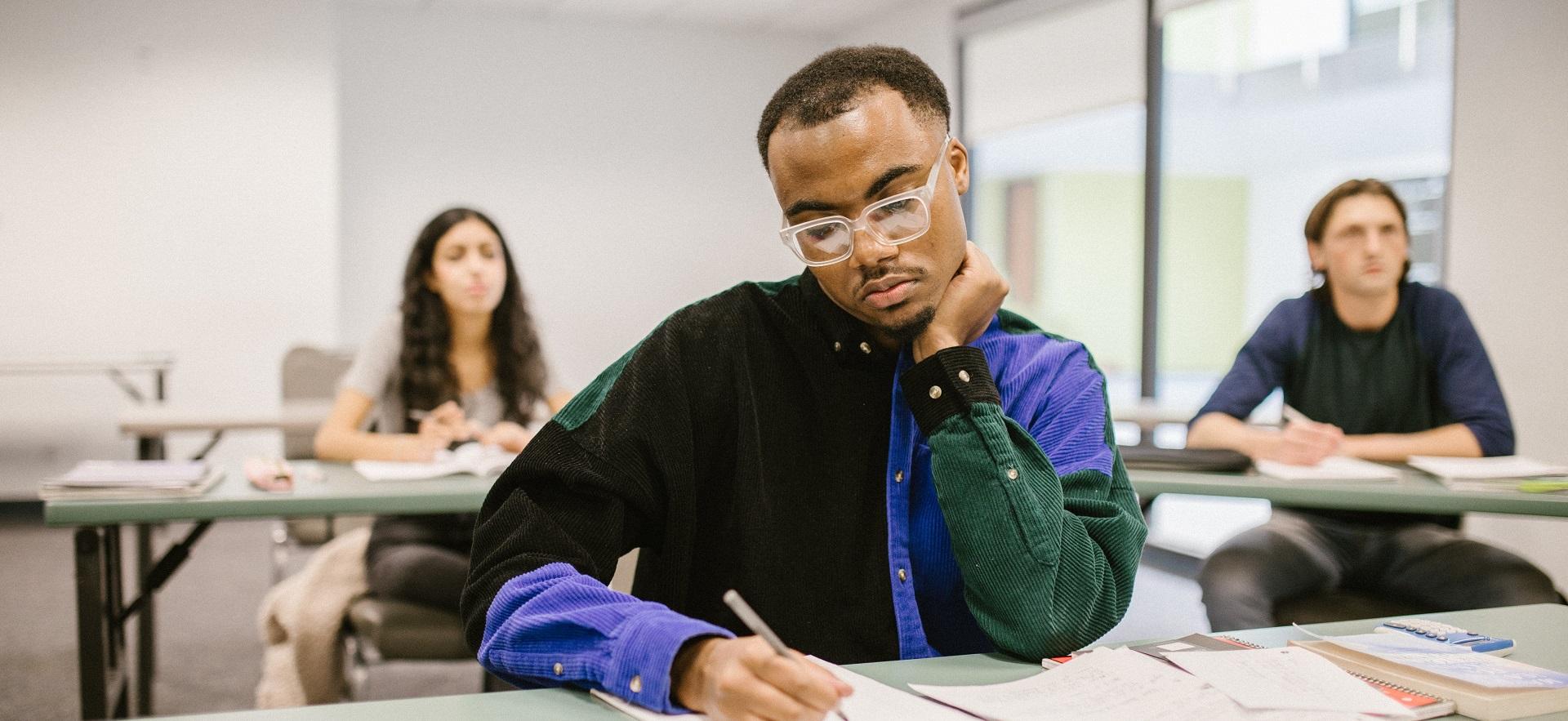 Student in classroom at desk looking down at paper with pen in hand and student in background