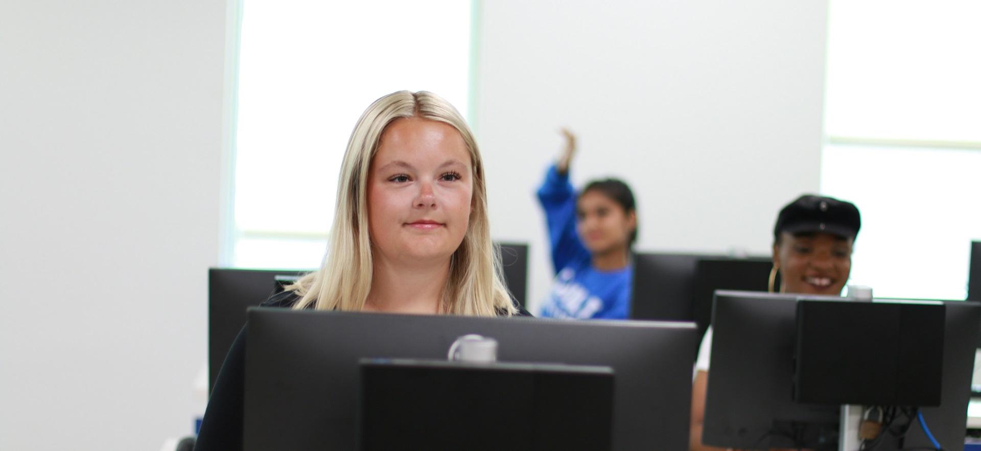 Female student at computer looking up at front of class with students at computers in background