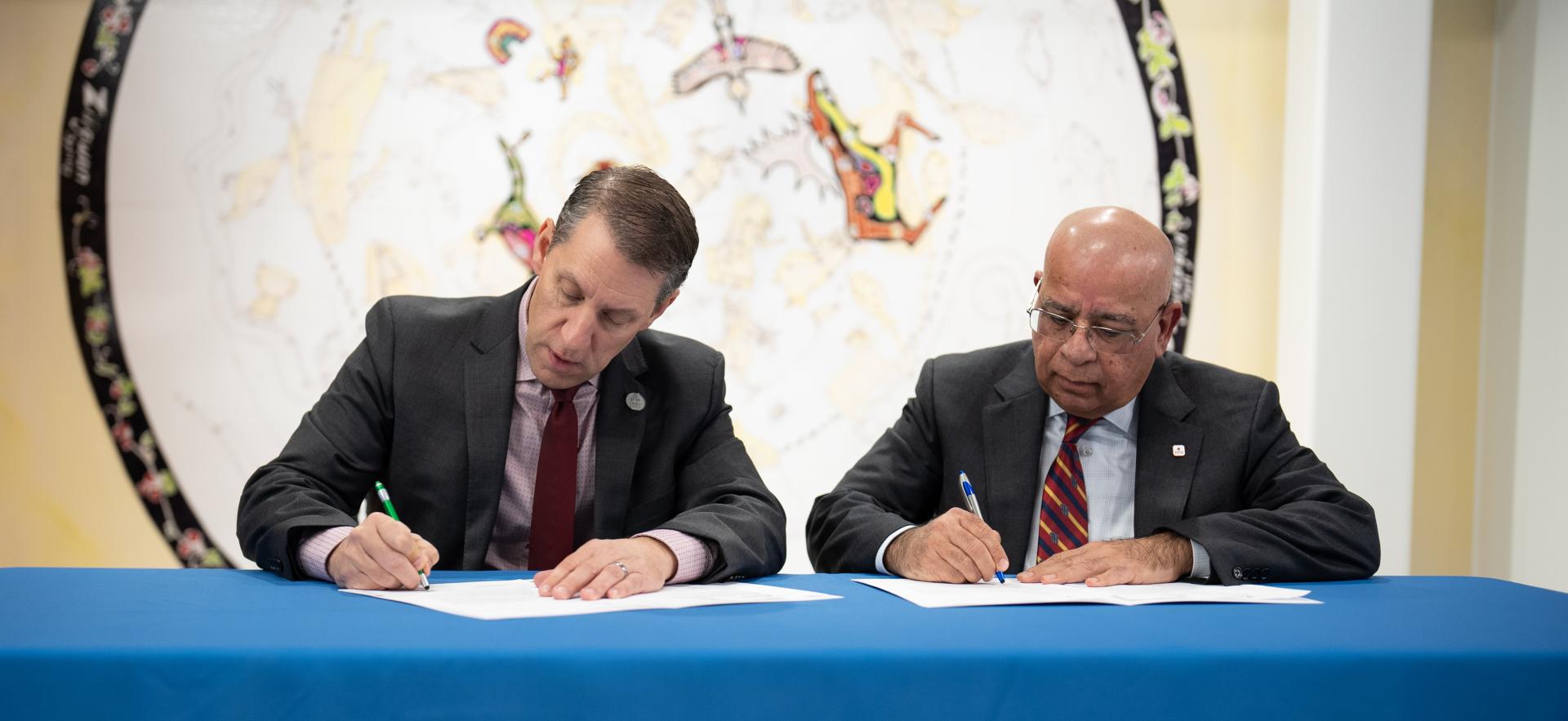two men in suits sitting at a table signing a paper. the tablecloth is blue