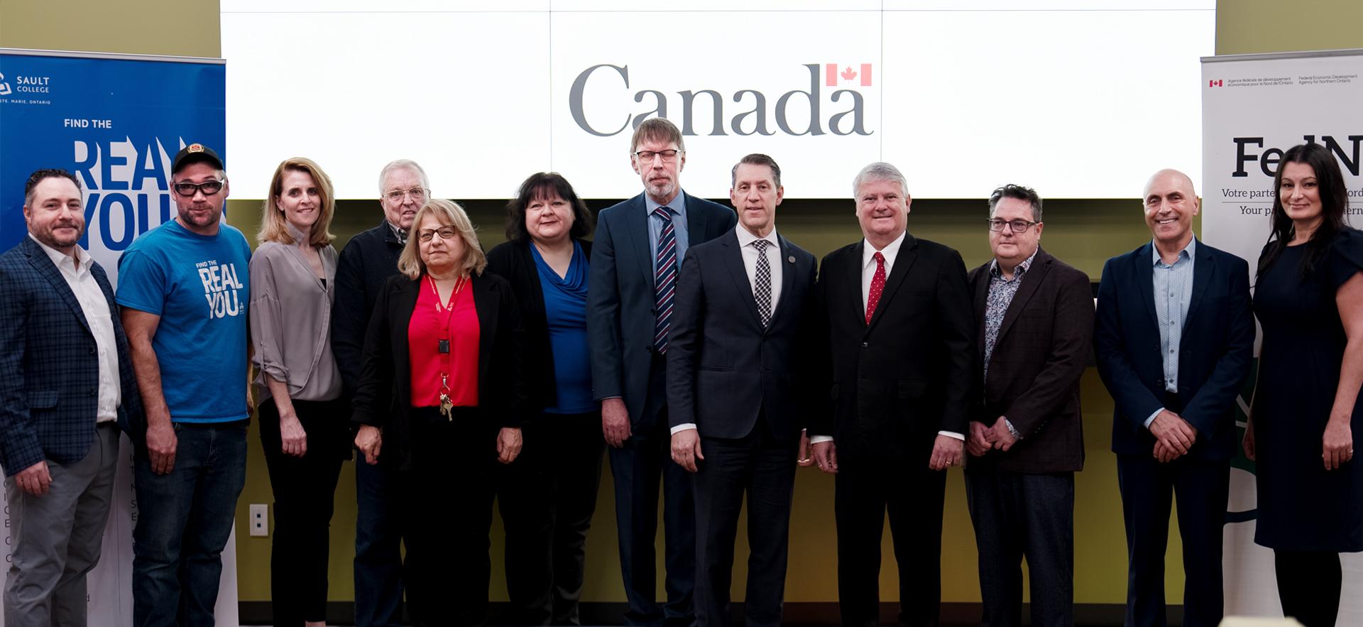 12 individuals standing in a row with the Government of Canada logo in the background