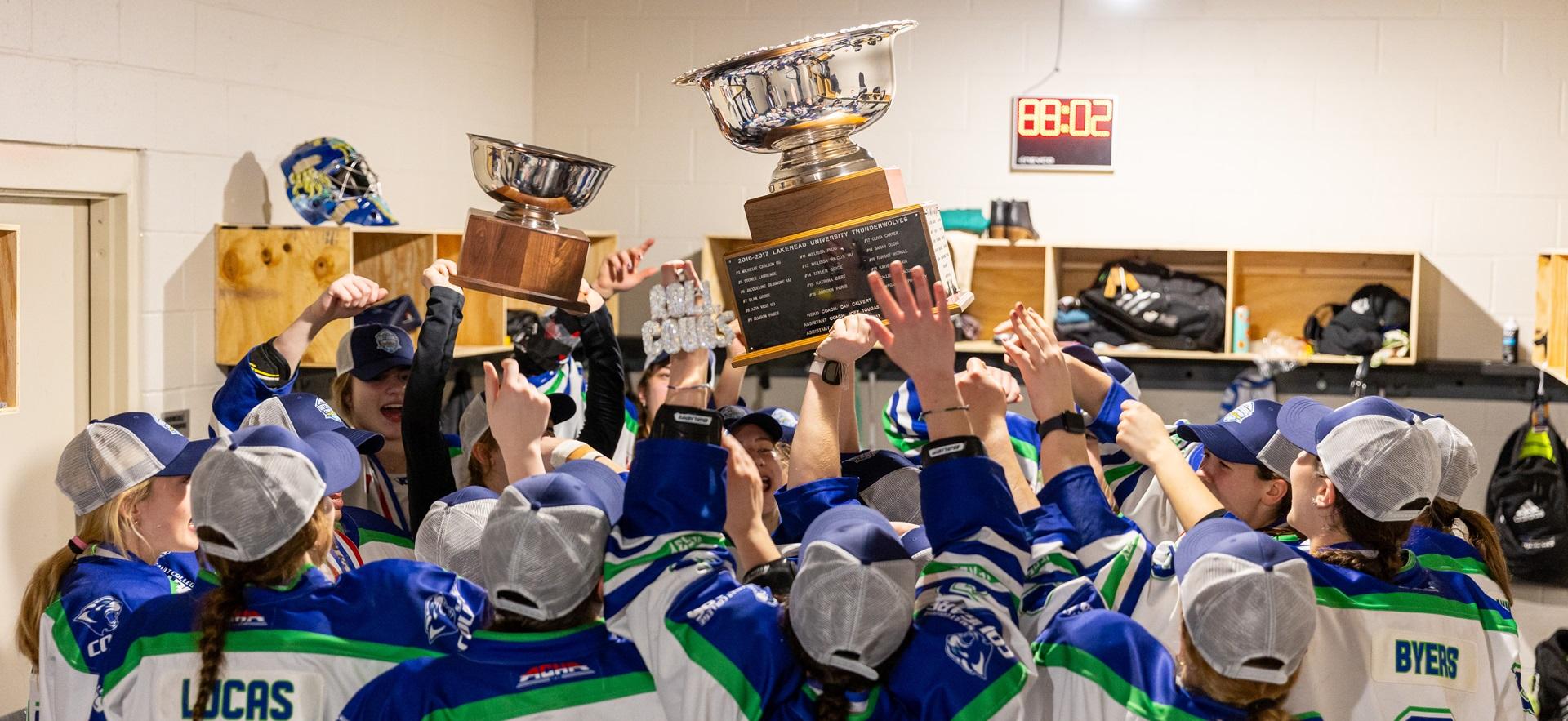 Women's hockey team gathered together in changeroom holding up trophies