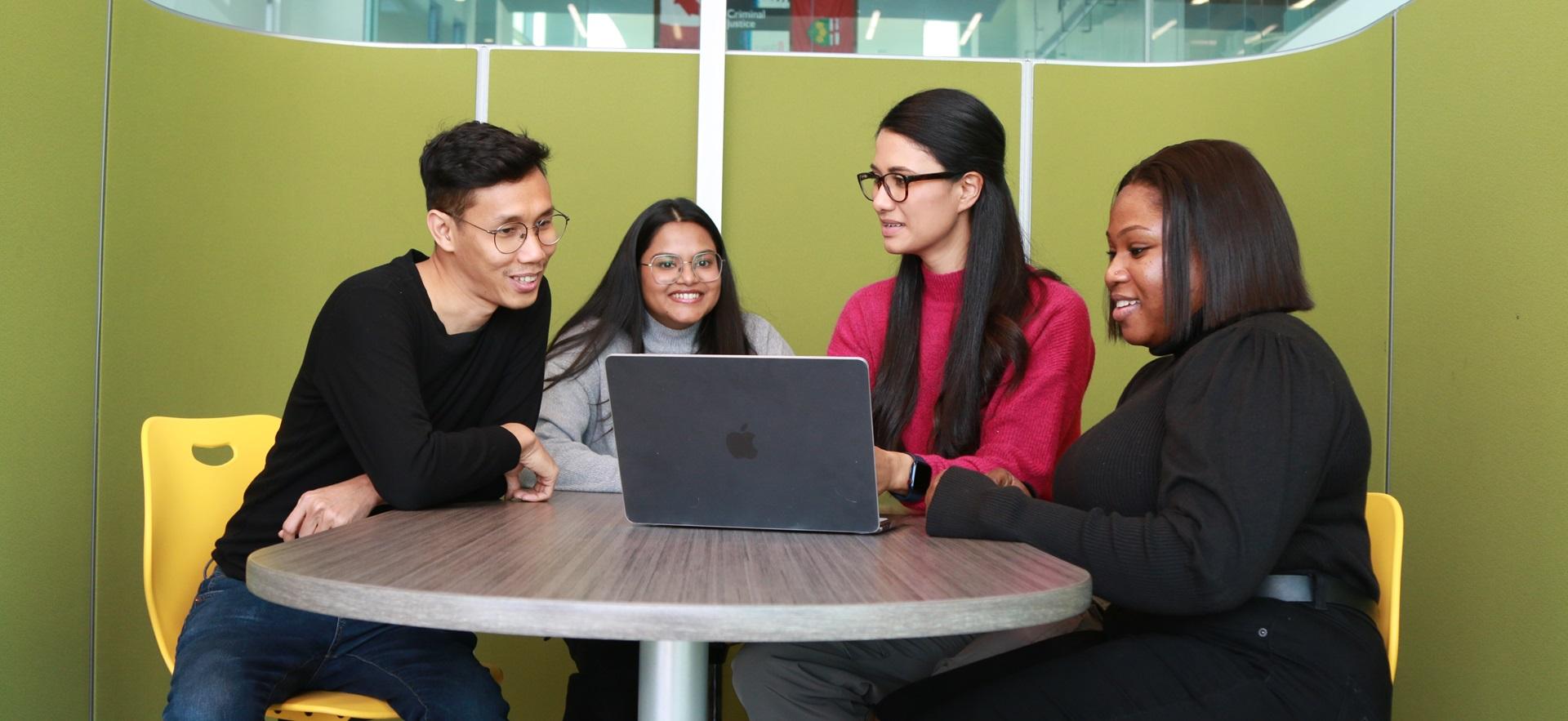 Four international students talking and smiling sitting around a table with a laptop