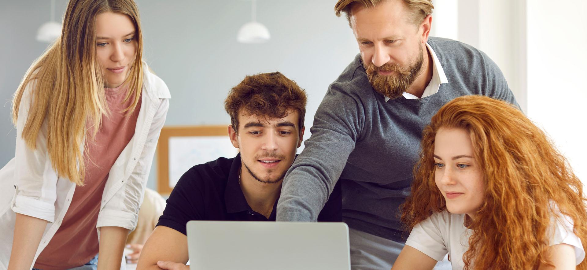 Adult man standing between two students sitting at a desk and one student standing on end