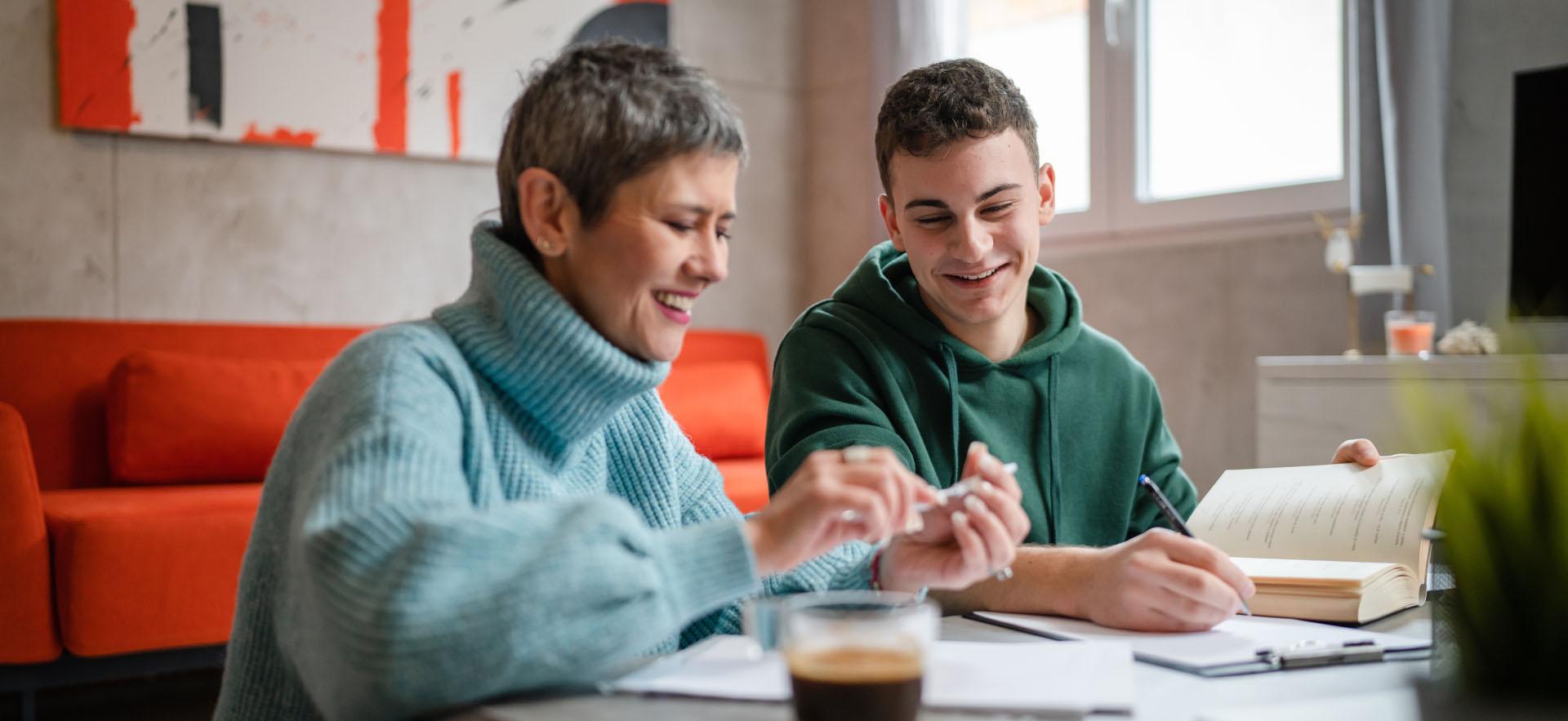 Adult woman sitting beside student at a desk smiling and looking down at papers