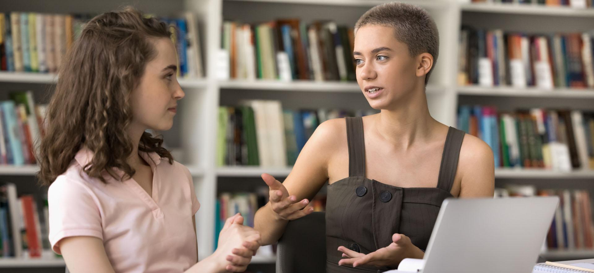 Two female students sitting together in library talking at a table