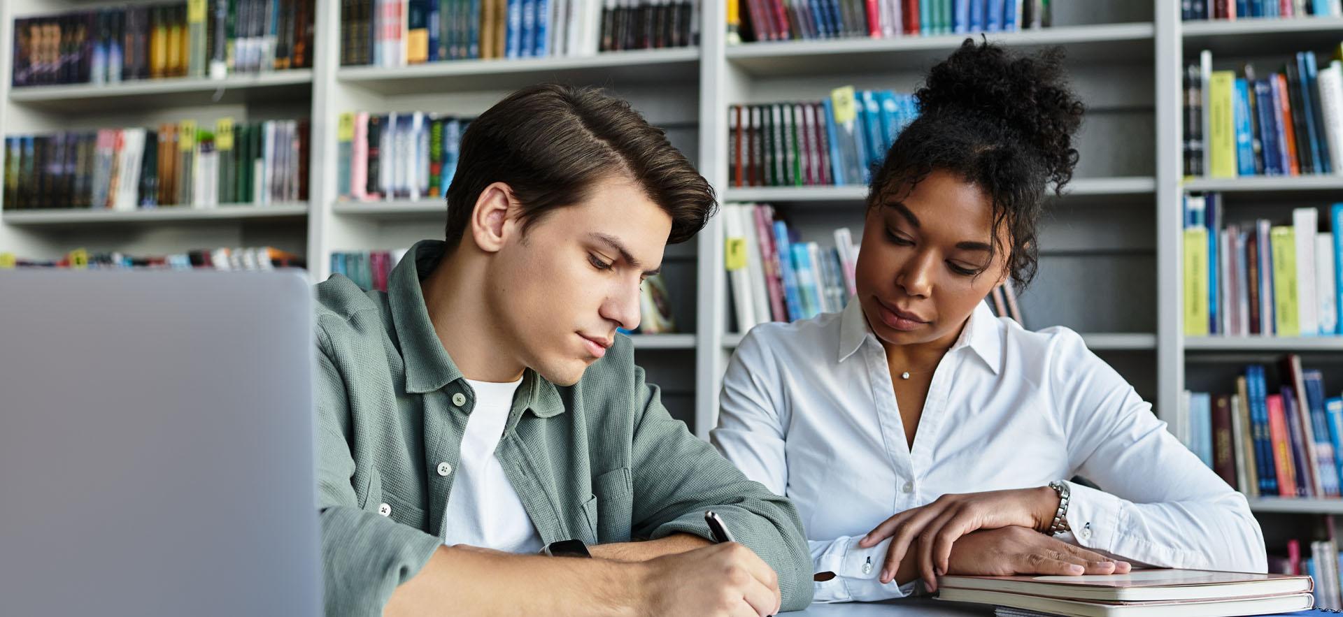 Two people sitting together looking at work on a table with bookshelf behind