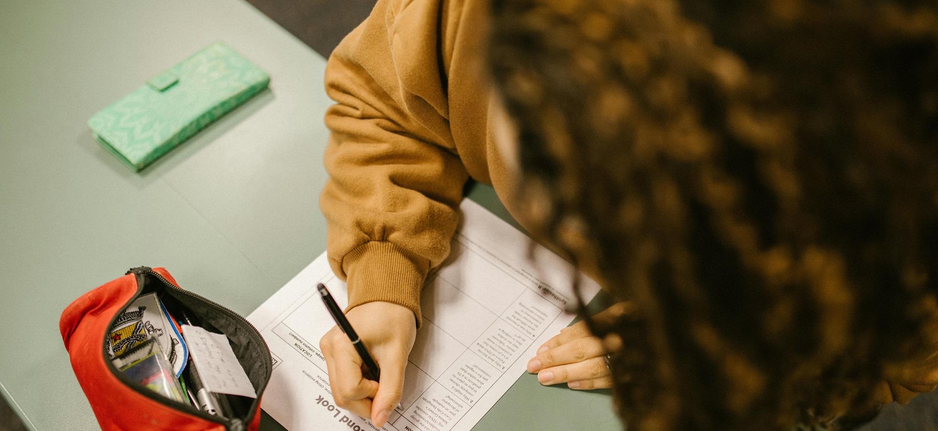 Student at desk looking down at paper with pen in hand writing