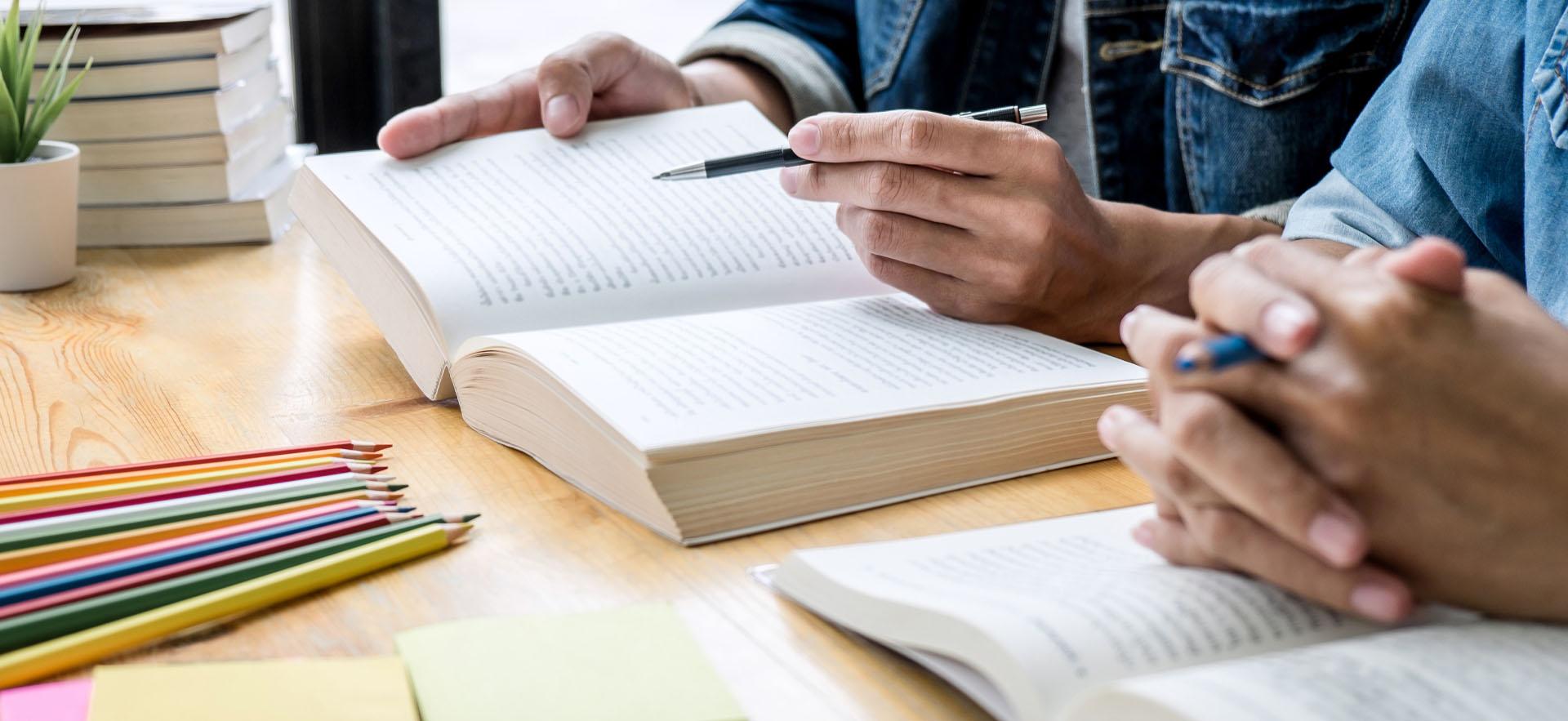Two students showing just their hands sitting at a table with books and a pen