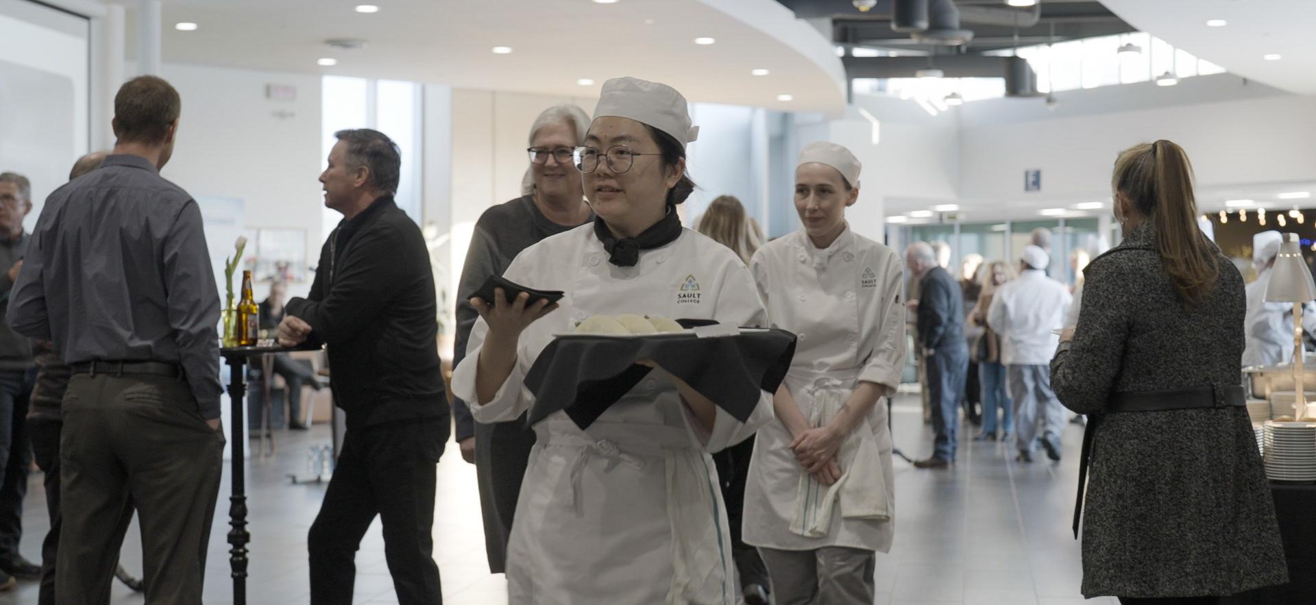 Culinary student shown carrying tray of hors d'oeuvres at event with people around