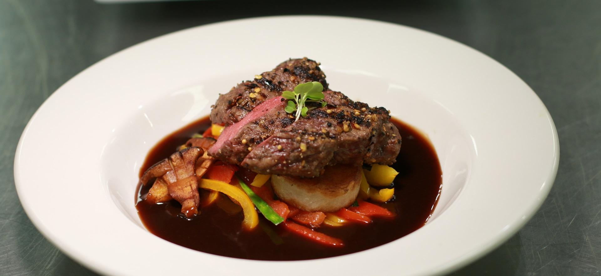 Culinary plated meal with a glaze in bowl around steak, vegetables and scallop