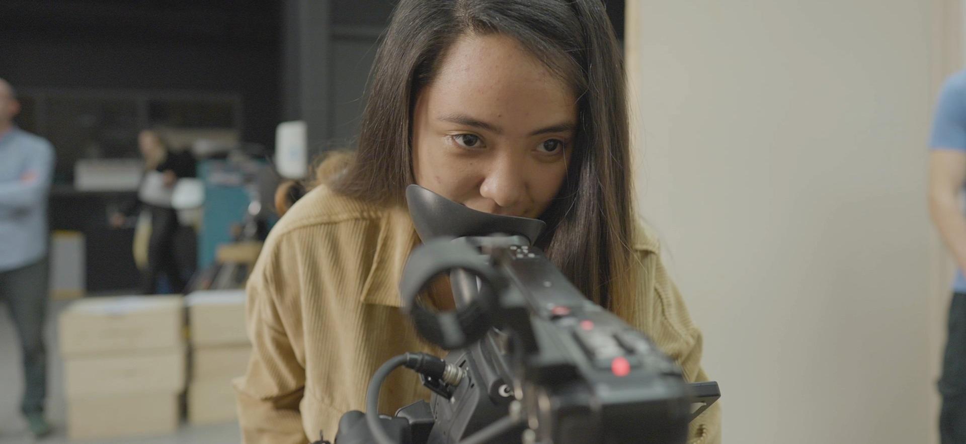 Film student shown face on leaning and looking at camera as she films on set