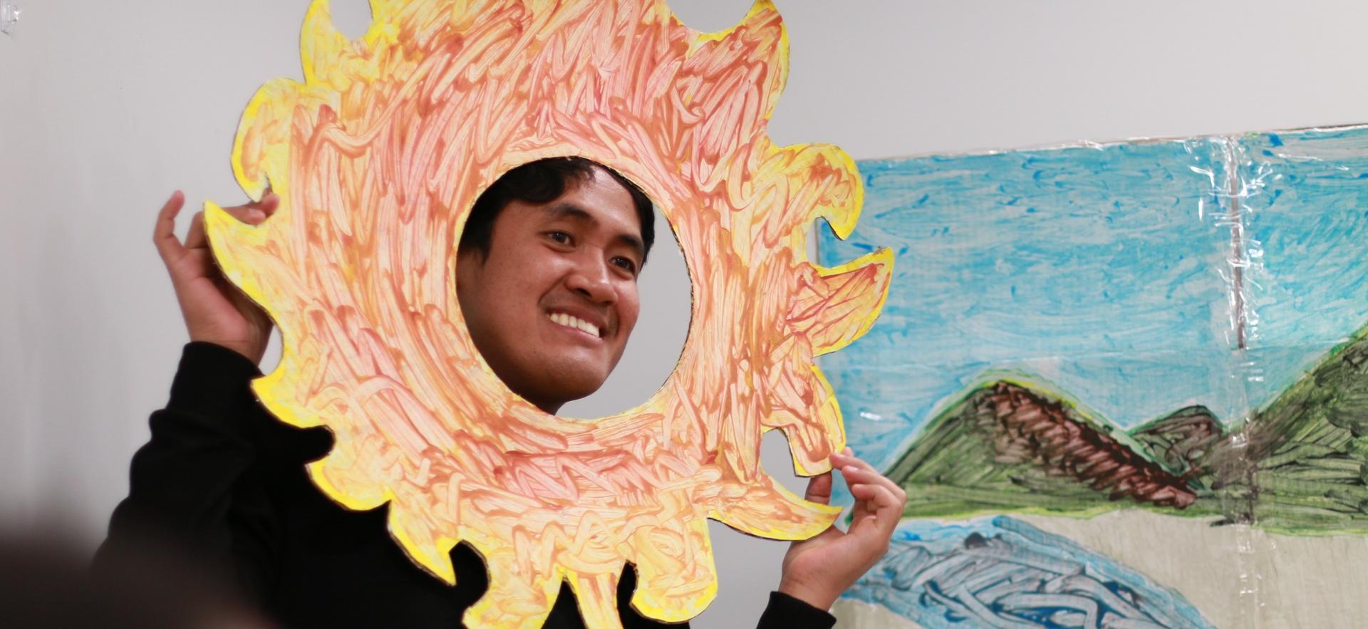 ECE student holding a large paper sun smiling and showing his face through a cut out