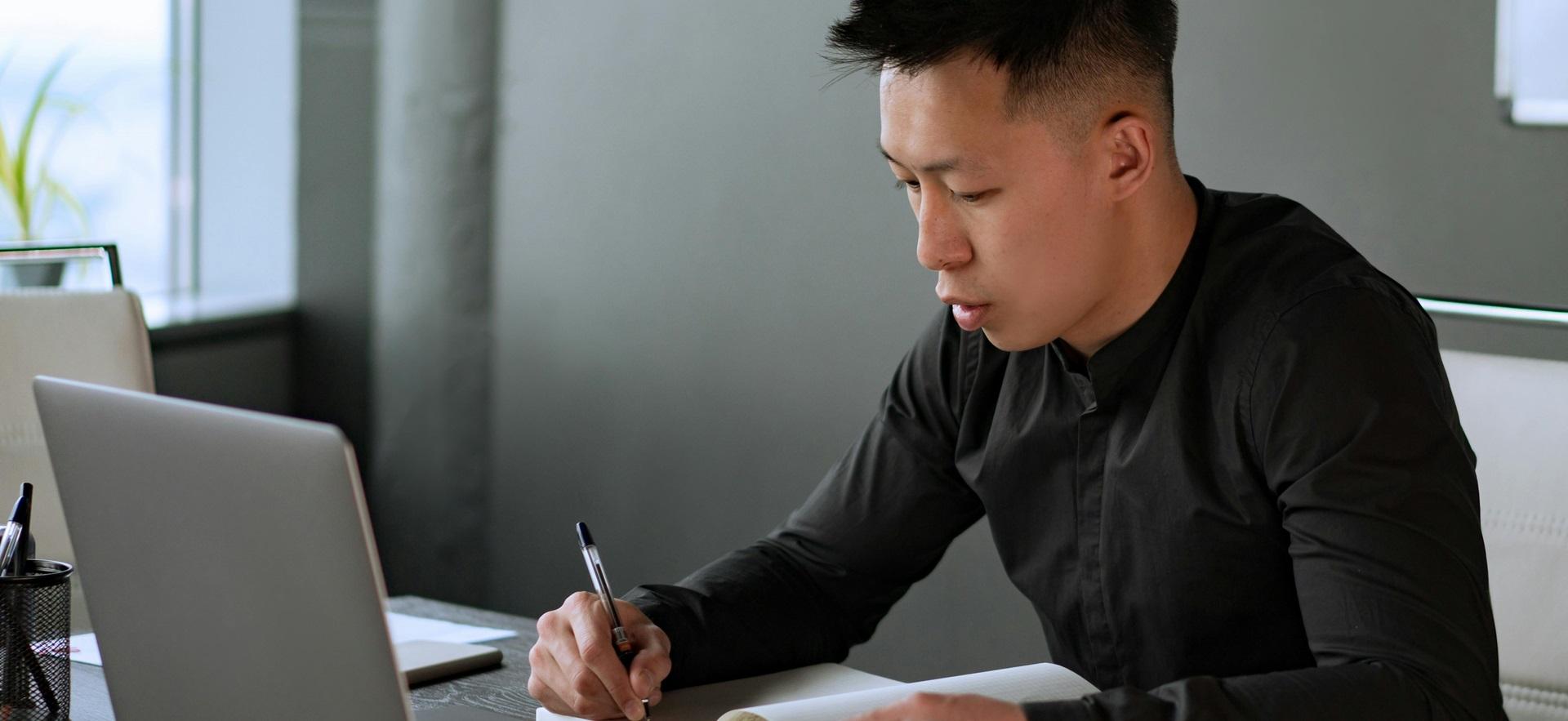 Man sitting at table at home with laptop and pen in hand writing in notebook