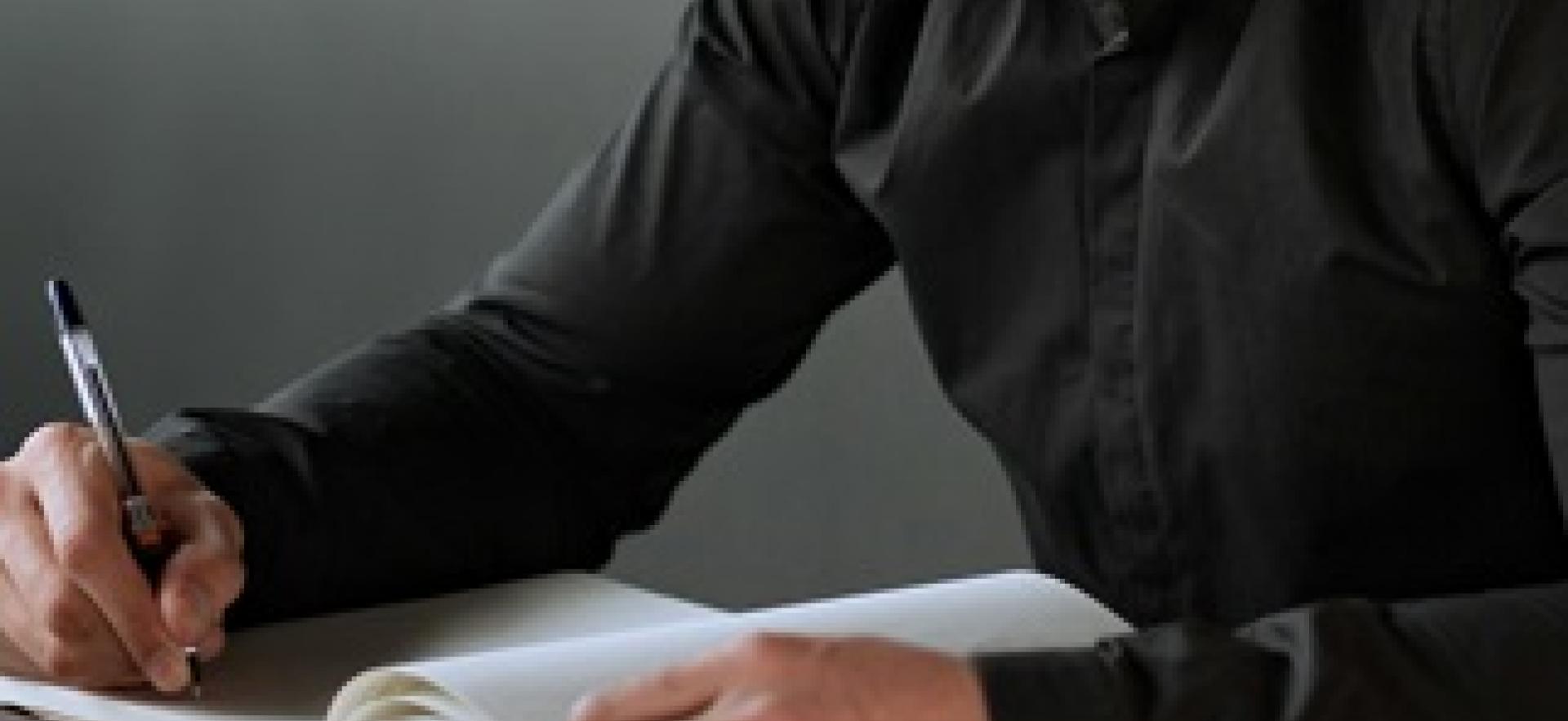 Man sitting at table at home with laptop and pen in hand writing in notebook